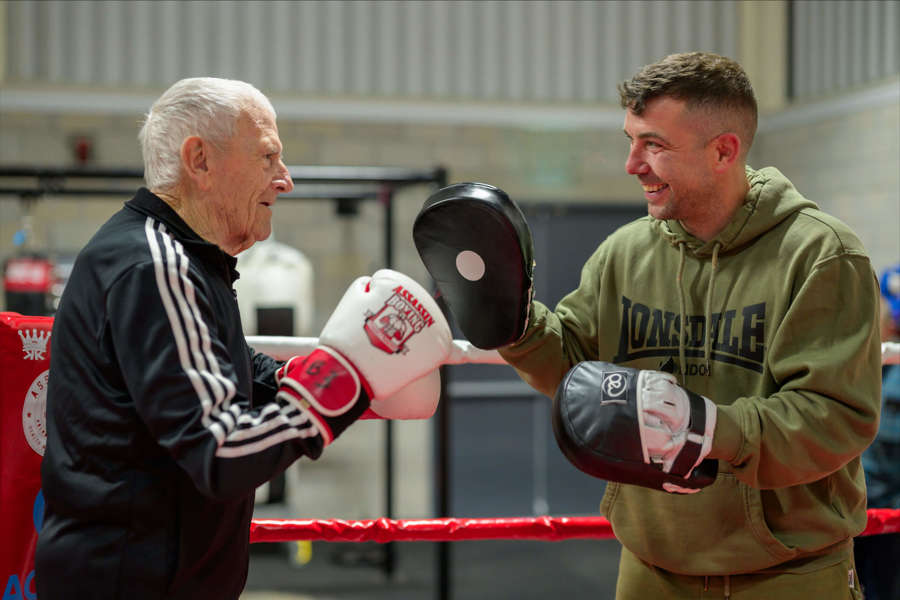 A care home resident and a local boxer face off in a boxing ring, both wearing gloves, ready for a friendly sparring match.