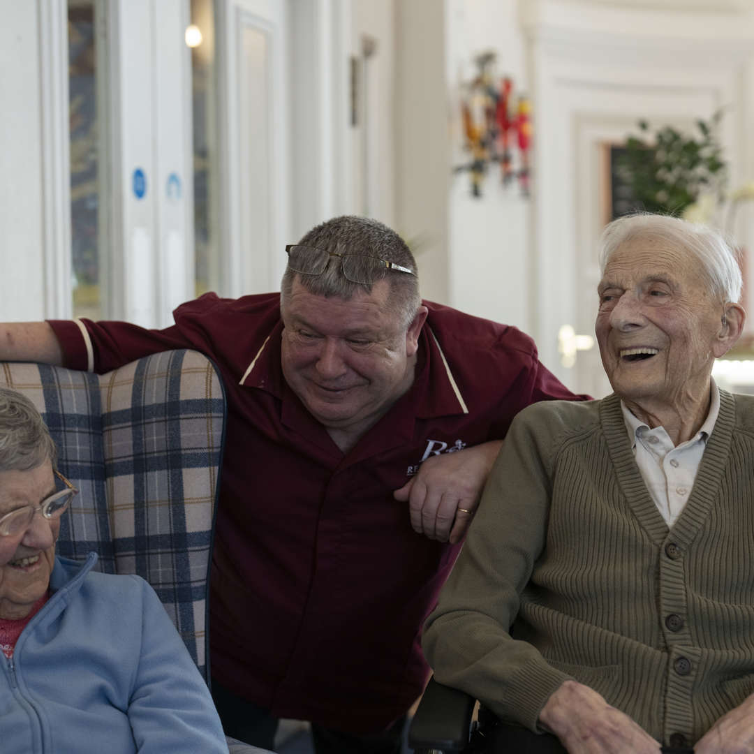 Care staff member attentively interacts with residents in a communal lounge. All are smiling and enjoying a friendly conversation