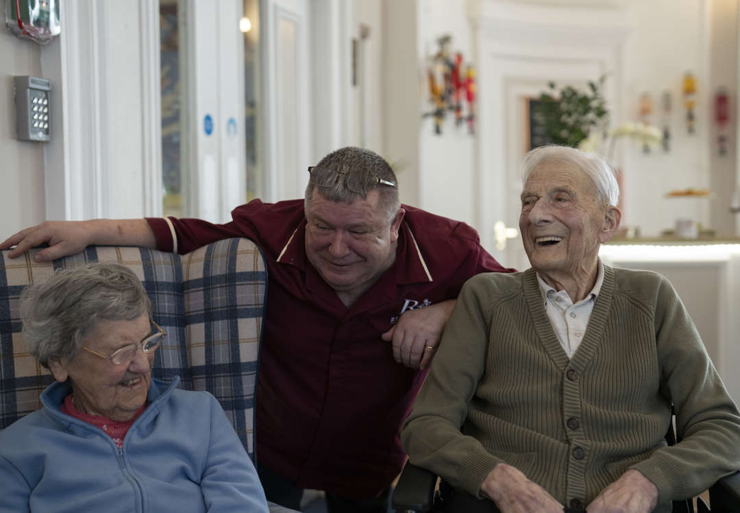 Care staff member attentively interacts with residents in a communal lounge. All are smiling and enjoying a friendly conversation