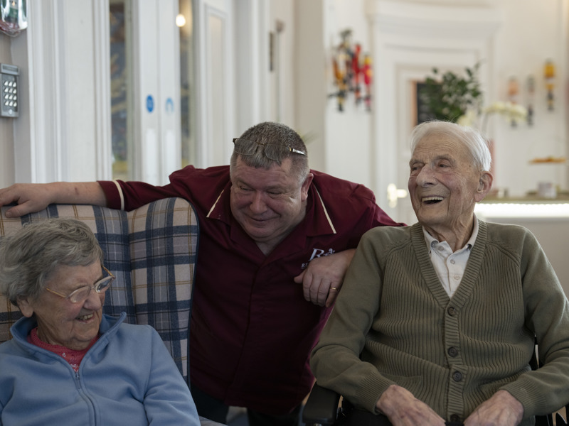 Care staff member attentively interacts with residents in a communal lounge. All are smiling and enjoying a friendly conversation