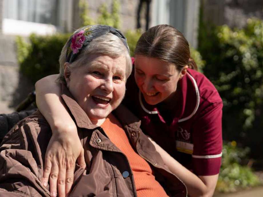 Caregiver offers a warm hug to a resident in a peaceful garden setting. The resident smiles, enjoying the moment of comfort and connection