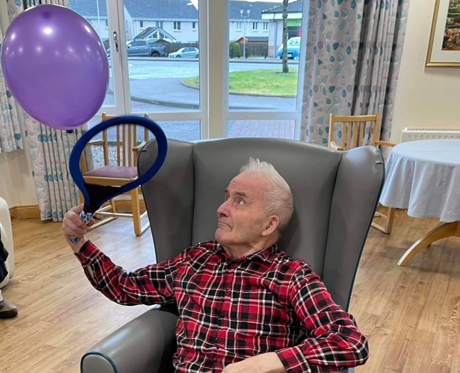 A care home resident sits in a high back chair. He is playing adapted tennis, with a racquet and a balloon