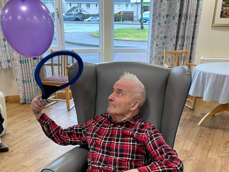 A care home resident sits in a high back chair. He is playing adapted tennis, with a racquet and a balloon