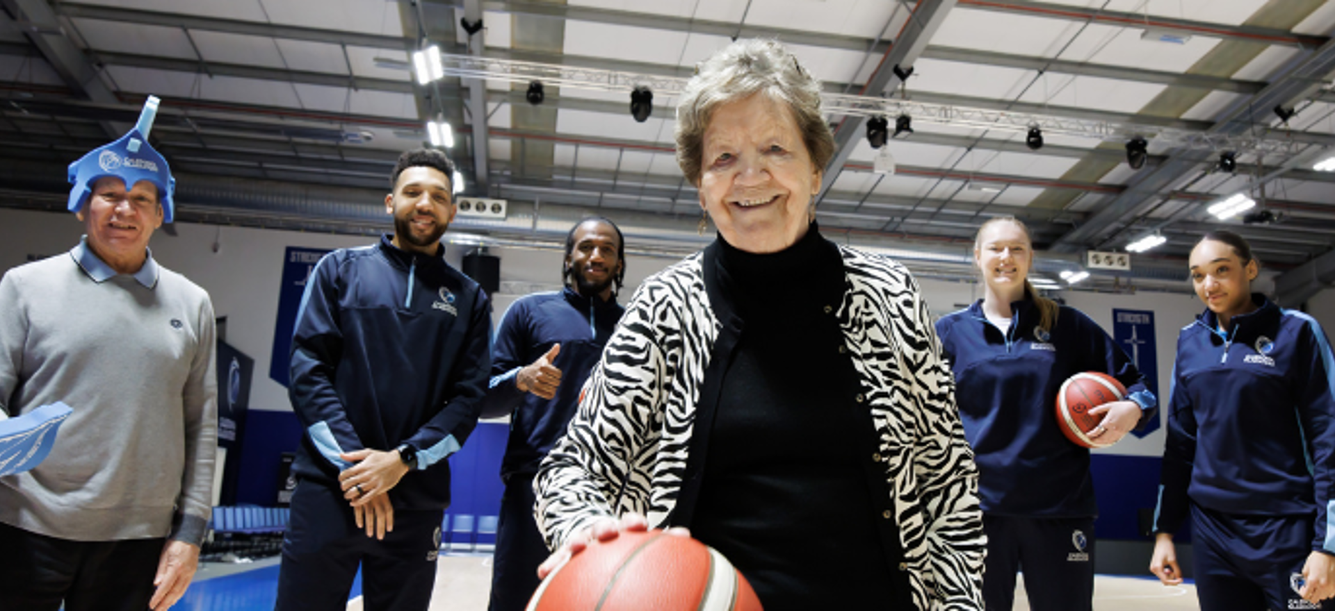 A care home resident at the forefront is bouncing a basketball, there are four Gladiator basketball players behind her and another care home resident with a foam finger and Gladiators headwear