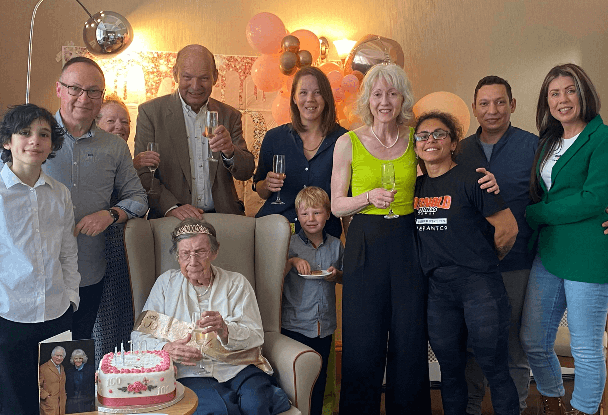 A care home resident is surrounded by her family at her 100th birthday celebrations. She is holding a prosecco. A side table holding her cake and card from the King sits in front of her. 