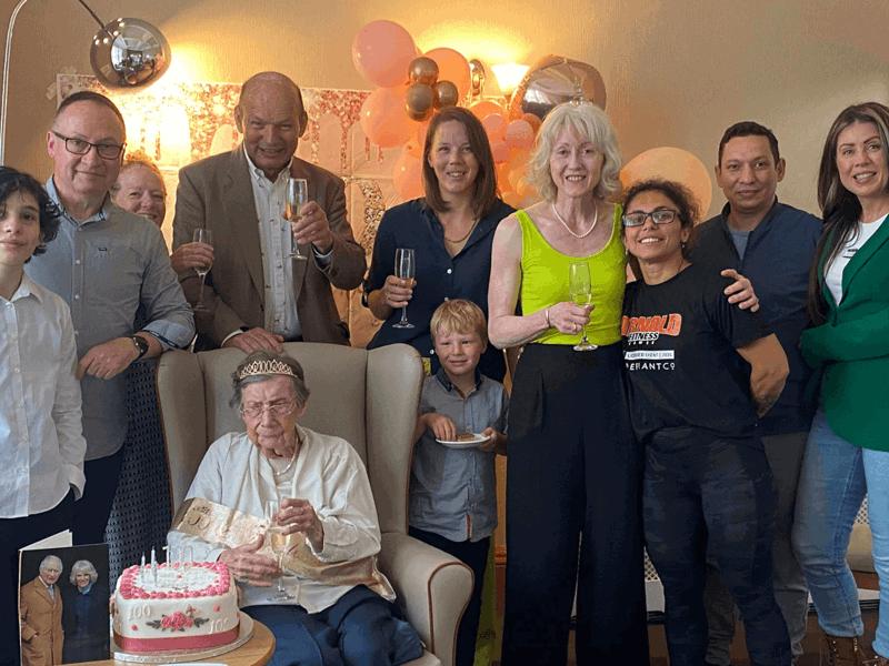 A care home resident is surrounded by her family at her 100th birthday celebrations. She is holding a prosecco. A side table holding her cake and card from the King sits in front of her. 