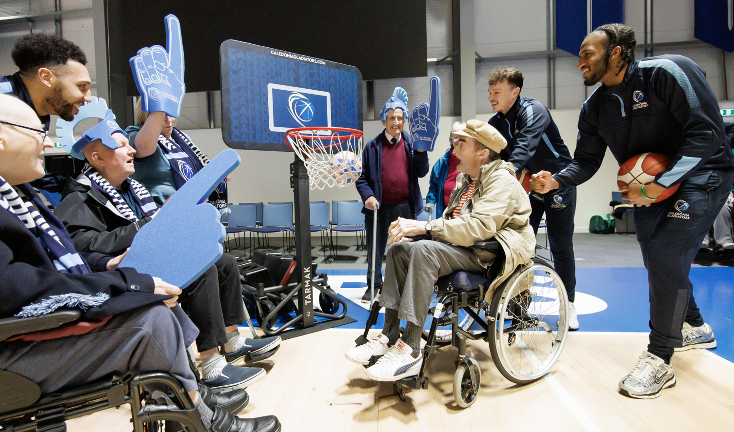 Residents and professional basketball players crowd around and cheer on care home resident Colin as he throws a basketball towards the hoop