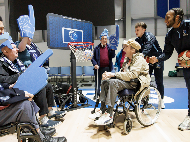 Residents and professional basketball players crowd around and cheer on care home resident Colin as he throws a basketball towards the hoop