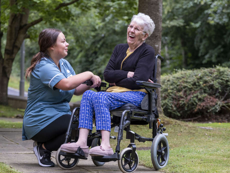 A staff member and resident are enjoying time in the garden. The staff member is kneeling down to the residents wheelchair, and they are smiling and laughing 