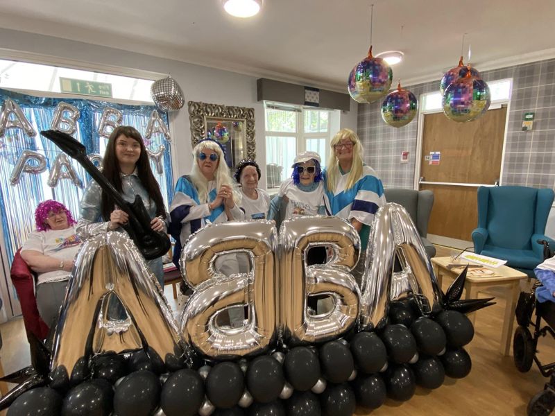 A group of care home residents and staff celebrating 50 years of ABBA with large balloons and themed decor