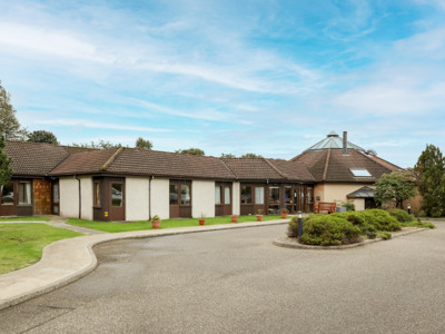 Meadowlark Care Home Exterior.  The building is white with brown accents around the windows and doors.  There is a large driveway or drop-off area in front of the building.  Trees and other greenery are planted around the building.