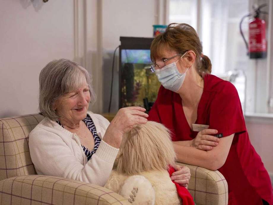 A woman sitting on an armchair within the homes communal area finds comfort with a teddy bear, as a staff member watches over her supportively.