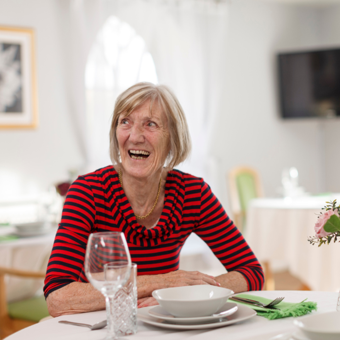 A woman sits at a dining room table, she is smiling 