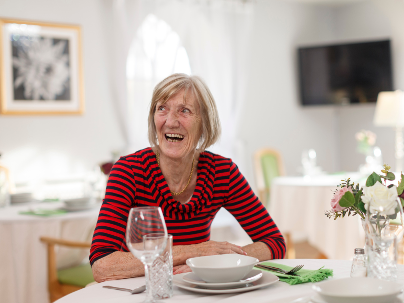 A woman sits at a dining room table, she is smiling 