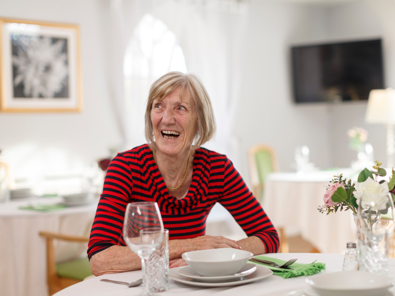 A woman sits at a dining room table, she is smiling