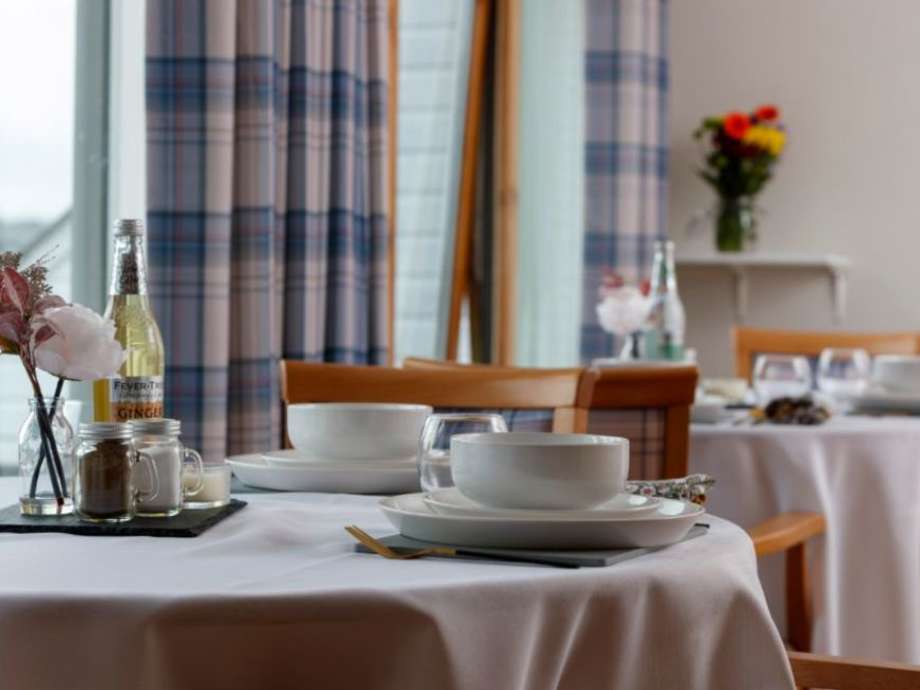 A dining room set up with a table and 4 chairs, napkins, plates, cutlery and flowers. There are windows with curtains to the rear 