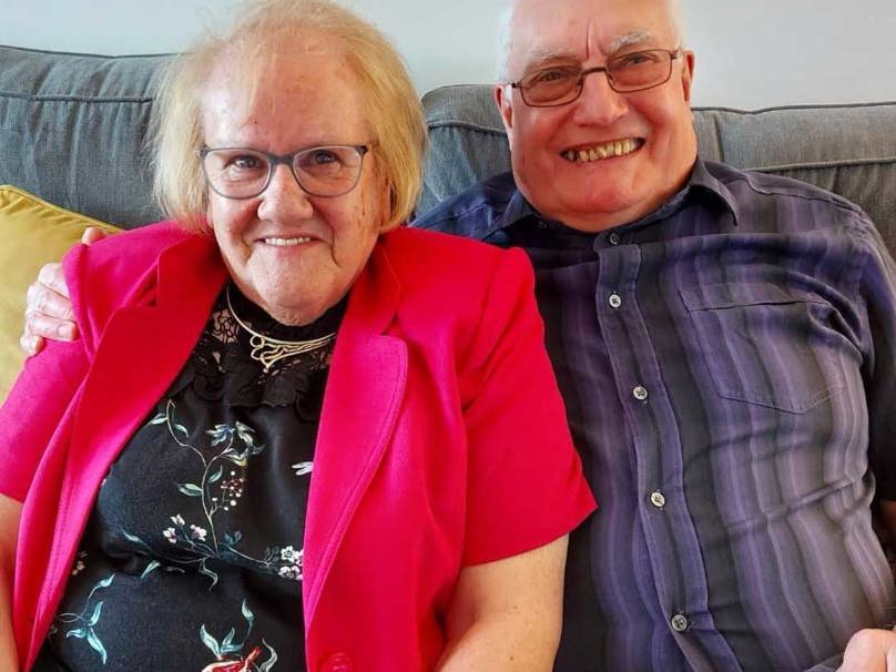 A happy couple celebrates Valentine's Day, sitting together on a sofa with big smiles.