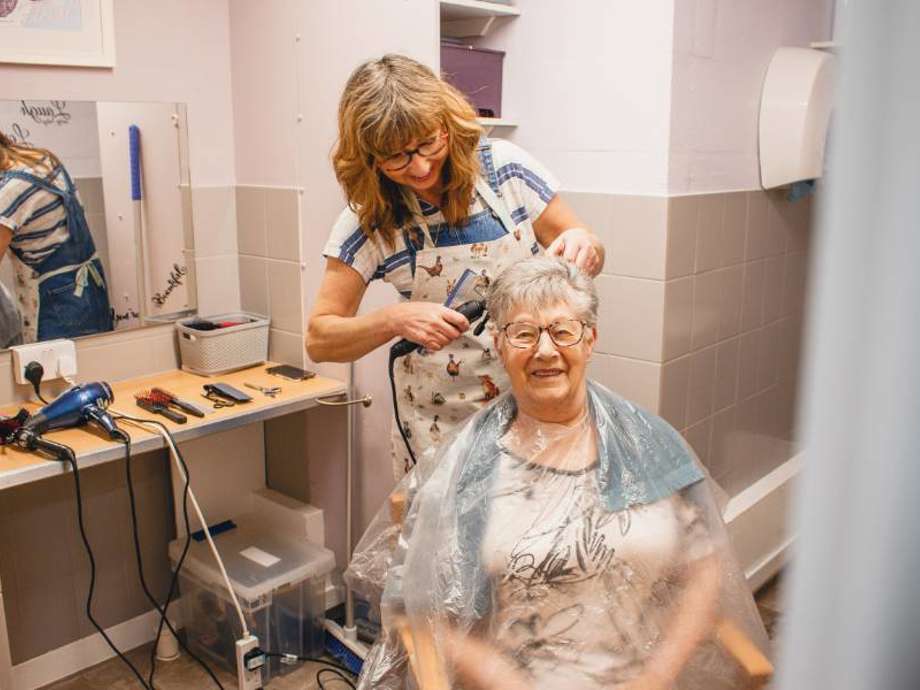 A care home resident enjoys a haircut from a visiting hairdresser. Both smile as the resident's hair is styled