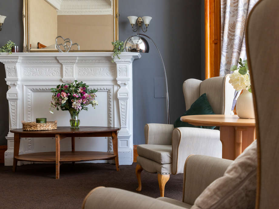 Milford House care home lounge: two high-packed chairs with a coffee table and a fireplace. Flowers and soft furnishings fill the room 