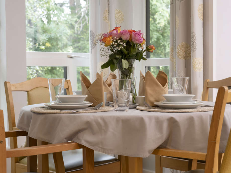 A dining room set up with a table and 4 chairs, napkins, plates, cutlery and fresh flowers. There are windows with curtains to the rear 