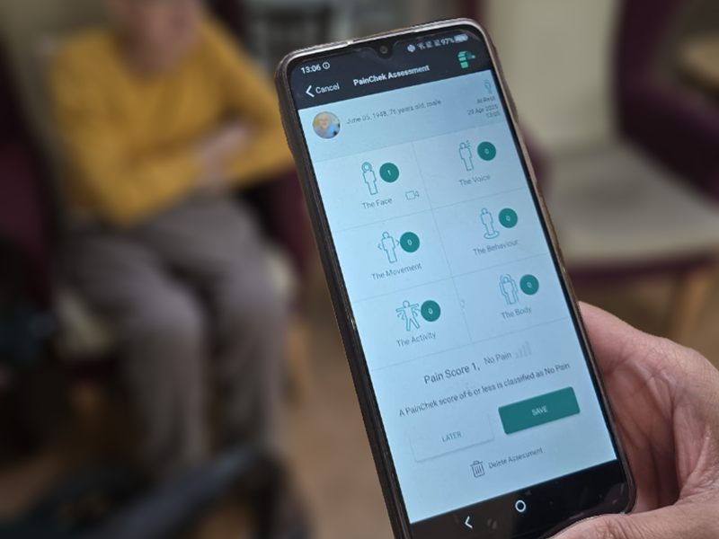A close up shot of a phone showing a PainChek pain assessment screen. The resident who's assessment is being completed sits comfortably on a high wingbacked armchair in the background.
