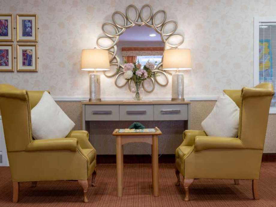 two yellow armchairs facing one another across a wooden table set for a game of Scrabble. A decorative sideboard with a mirror reflects a vase of flowers and elegant lamps.