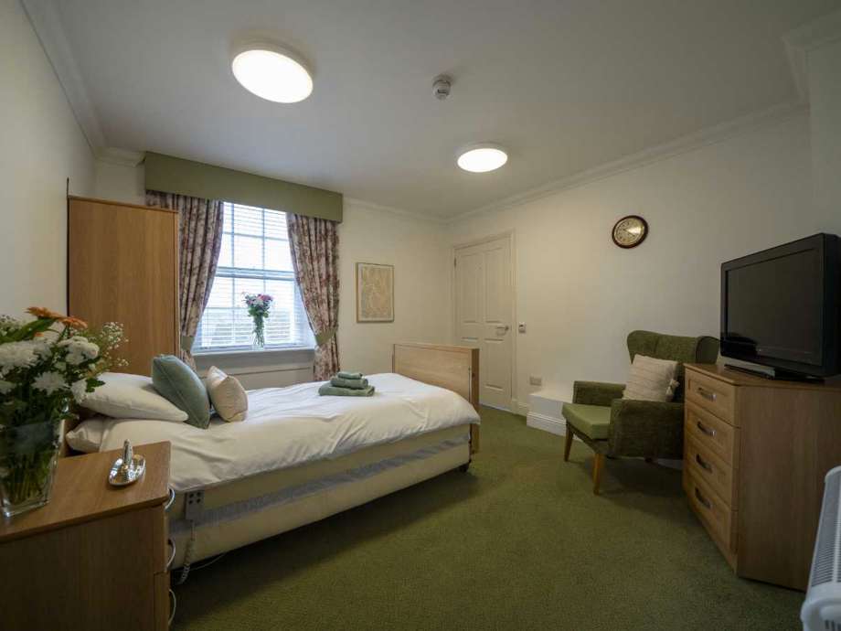 A tranquil care home bedroom with inviting sage green accents, featuring a comfortable bed, bedside unit, TV unit, and chair.