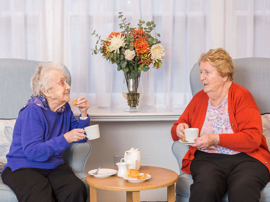 Two residents sit on high backed armchairs enjoying a cup of tea together and a chat