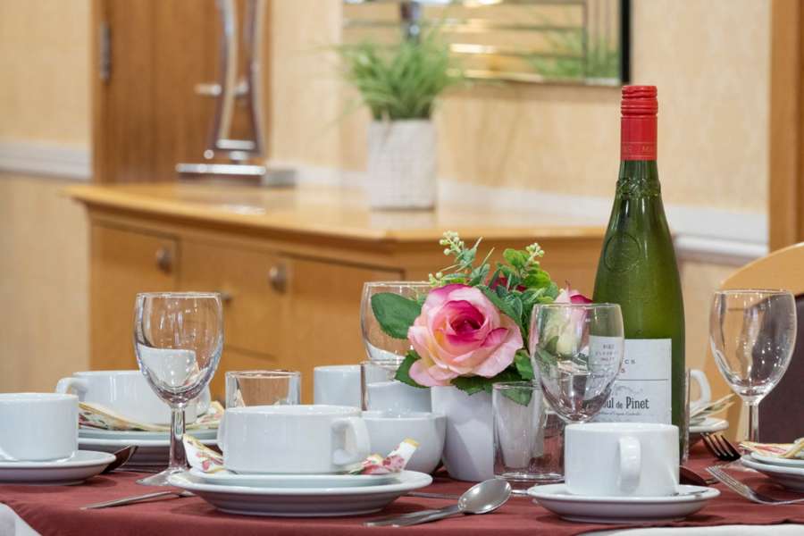 A decorative table set up in the dining room. A table with a red tablecloth and an array of teacups, wine glasses, flowers & a bottle of wine