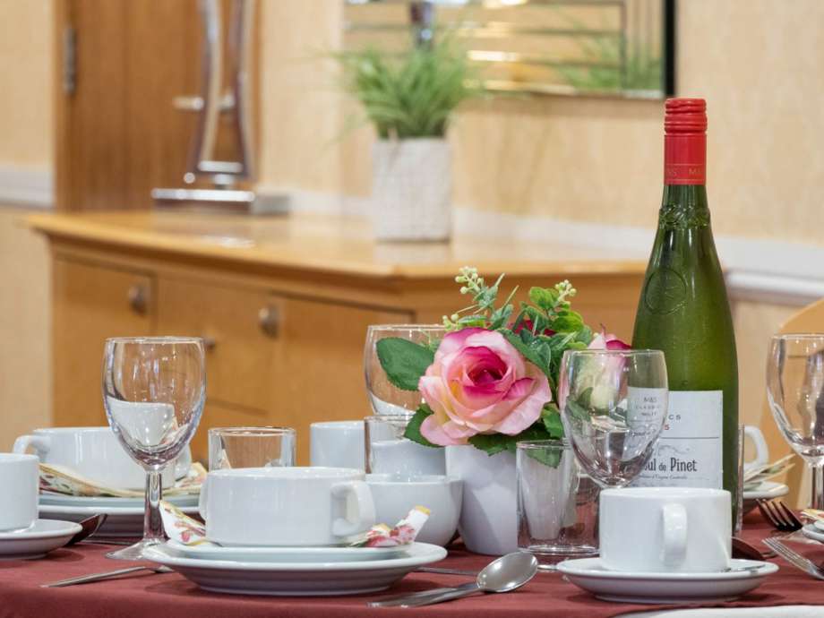 A decorative table set up in the dining room. A table with a red tablecloth and an array of teacups, wine glasses, flowers & a bottle of wine