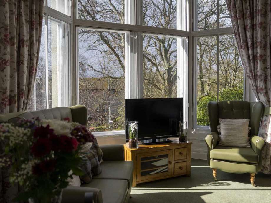 Comfortable care home bedroom lounge featuring a couch, armchair, and TV unit for relaxation. Large windows offer a scenic view of the garden below