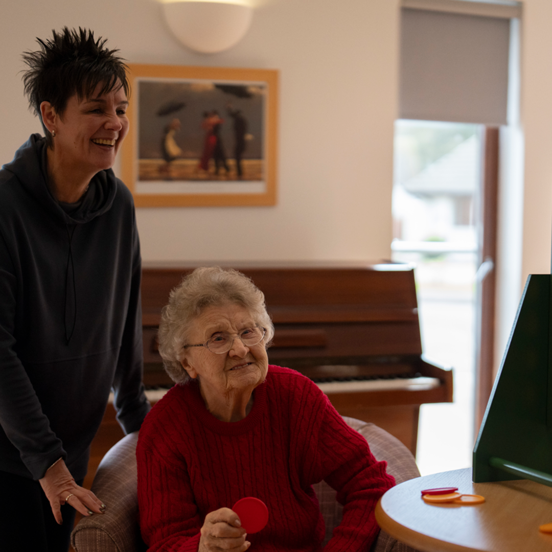 two woman are playing a game of connect 4. There is a large game board on the table next to them, with one of the women sat and the other stood beside it