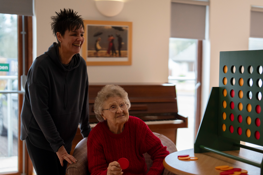 two woman are playing a game of connect 4. There is a large game board on the table next to them, with one of the women sat and the other stood beside it