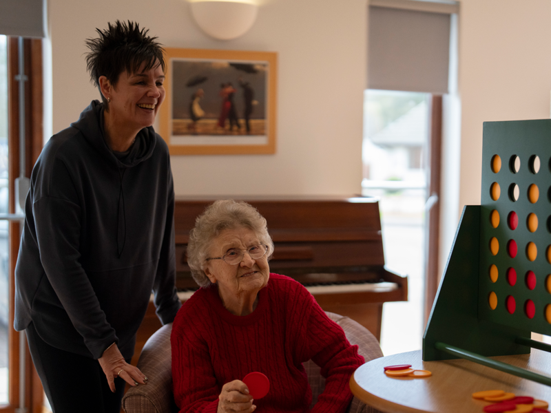 two woman are playing a game of connect 4. There is a large game board on the table next to them, with one of the women sat and the other stood beside it