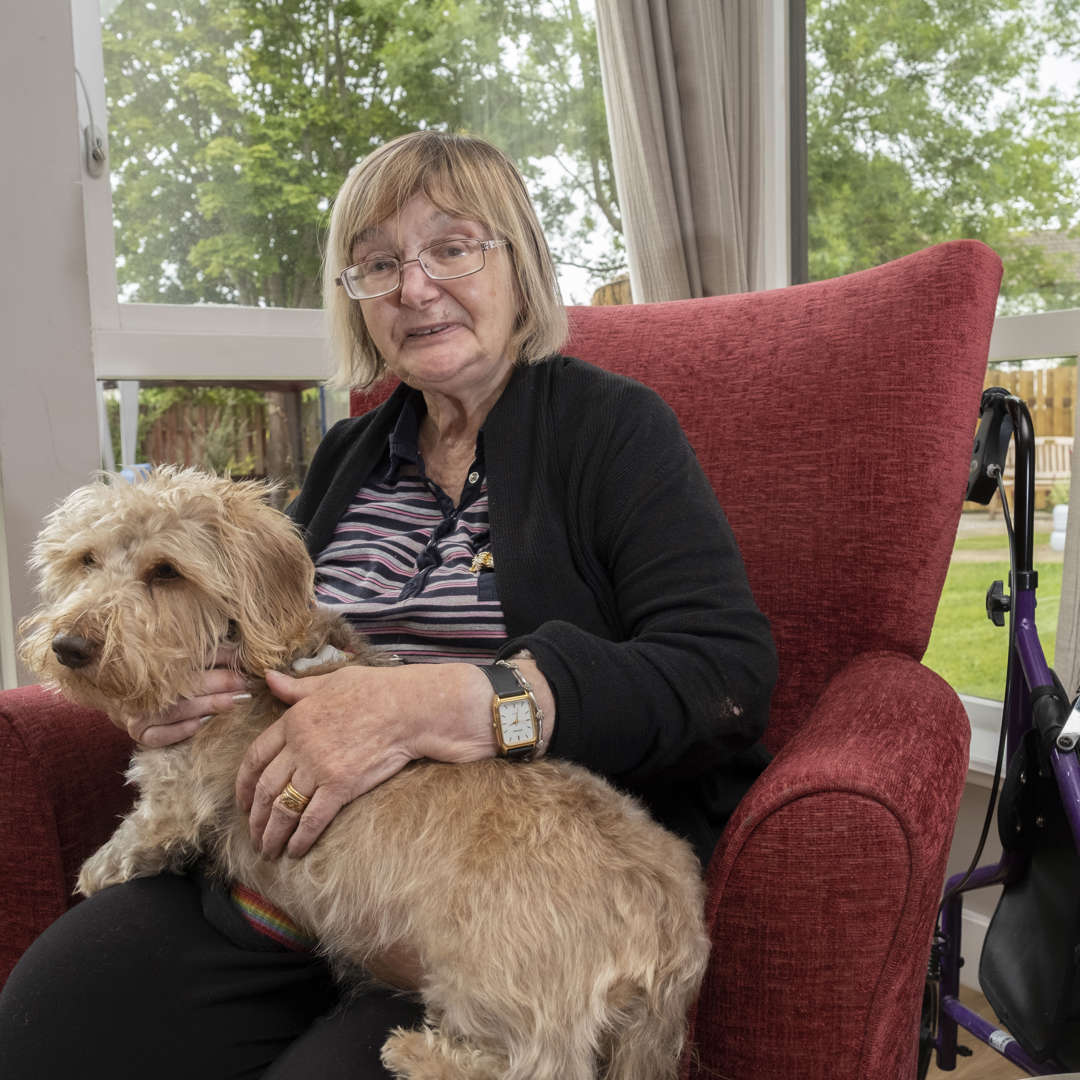 A woman sits on a red armchair next to the window looking out to the garden. She is enjoying a cuddle from therapet, Dexter the dog