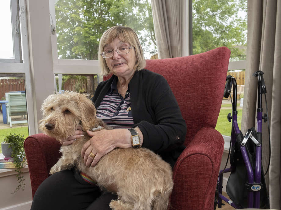 A woman sits on a red armchair next to the window looking out to the garden. She is enjoying a cuddle from therapet, Dexter the dog