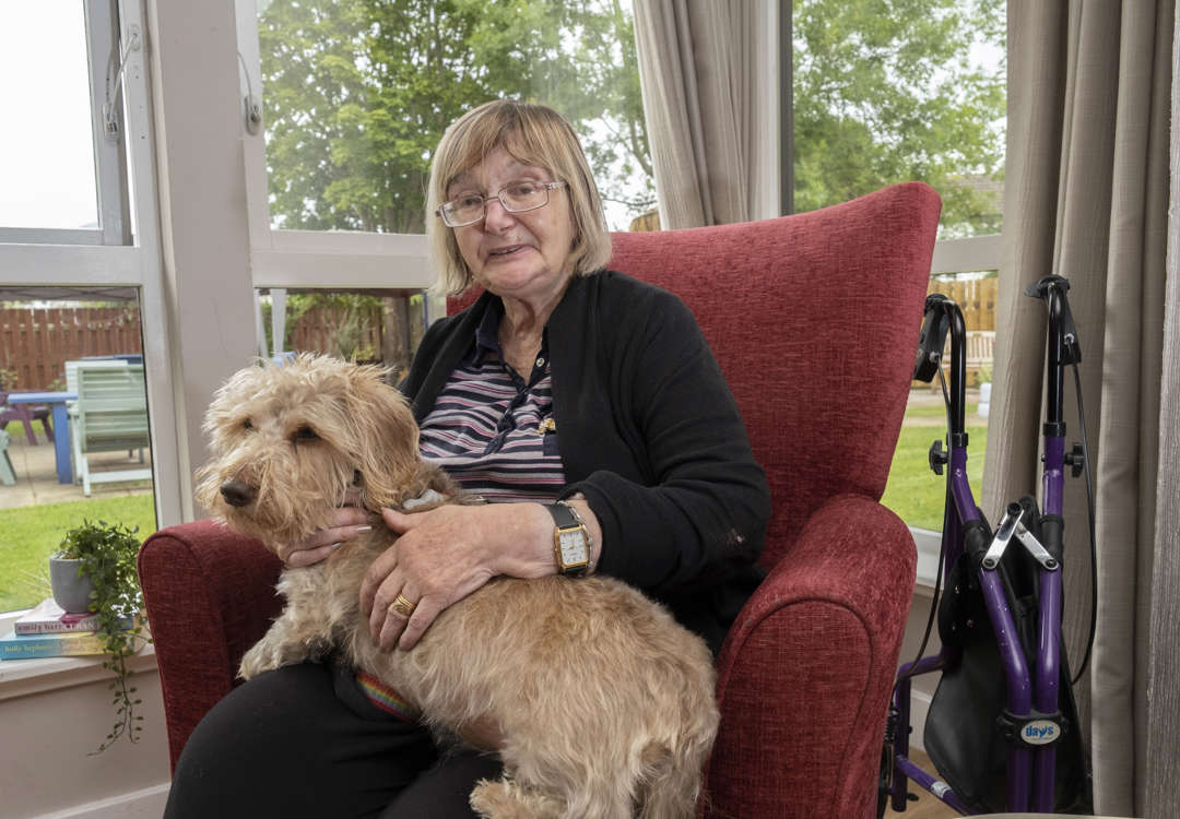 A woman sits on a red armchair next to the window looking out to the garden. She is enjoying a cuddle from therapet, Dexter the dog