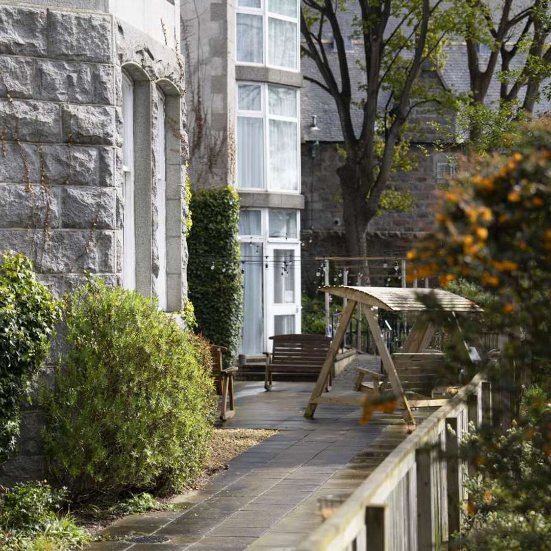 Serene care home garden featuring a relaxing patio with comfortable seating. Lush greenery surrounds the space, creating a tranquil atmosphere.