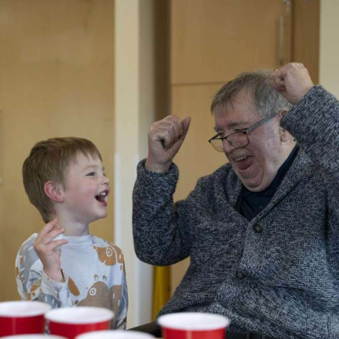 A care home resident smiles and cheers while playing a ball and cup game with a visiting nursery child