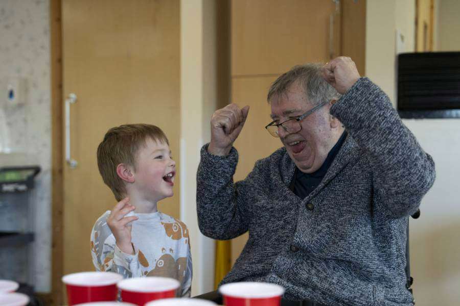 A care home resident smiles and cheers while playing a ball and cup game with a visiting nursery child