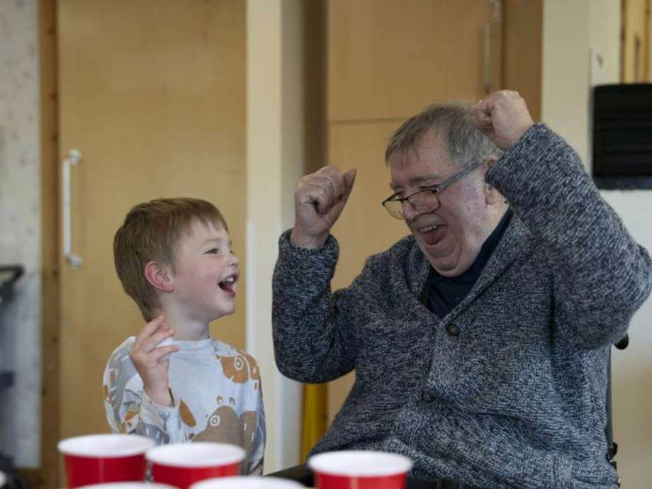 A care home resident smiles and cheers while playing a ball and cup game with a visiting nursery child