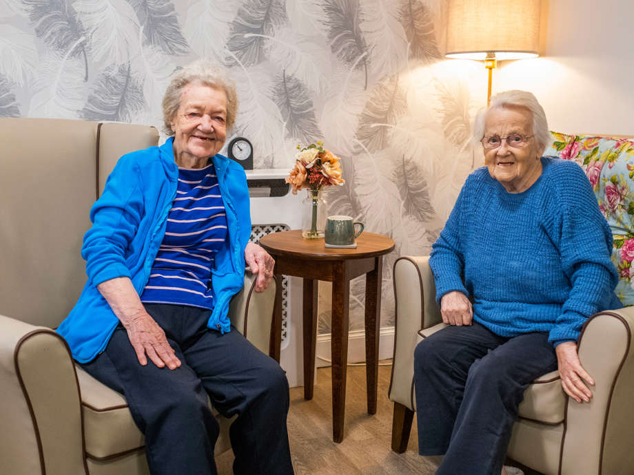 Two residents, wearing blue, share a warm conversation and smiles in plush armchairs