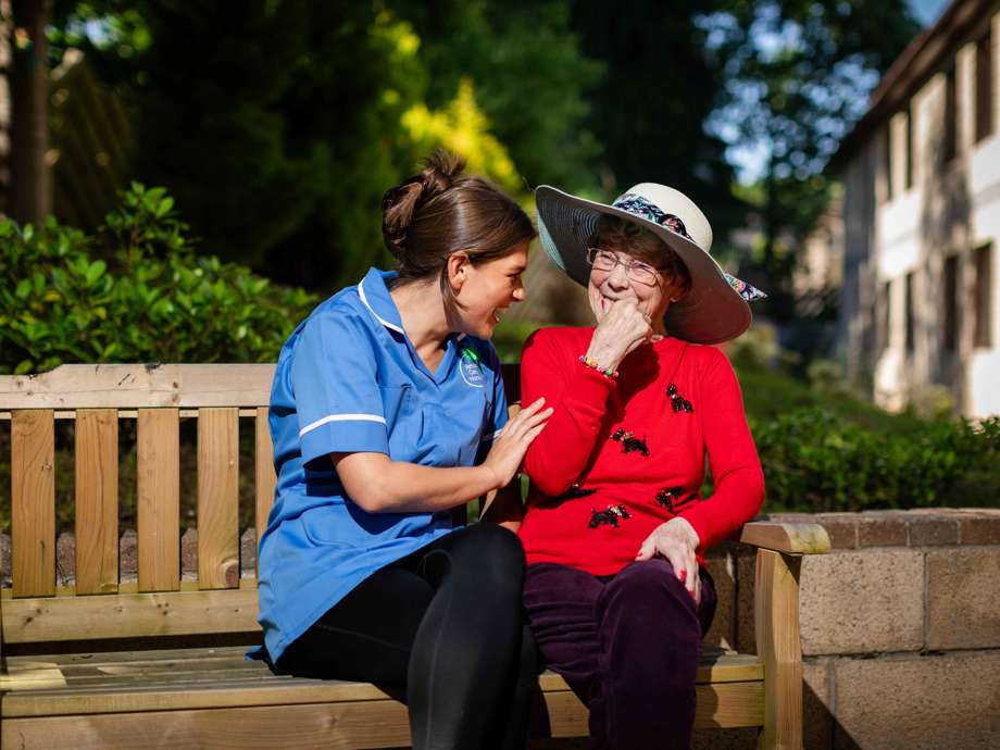 A resident and staff member enjoy one-on-one time outdoors. They sit on a bench in the homes garden area