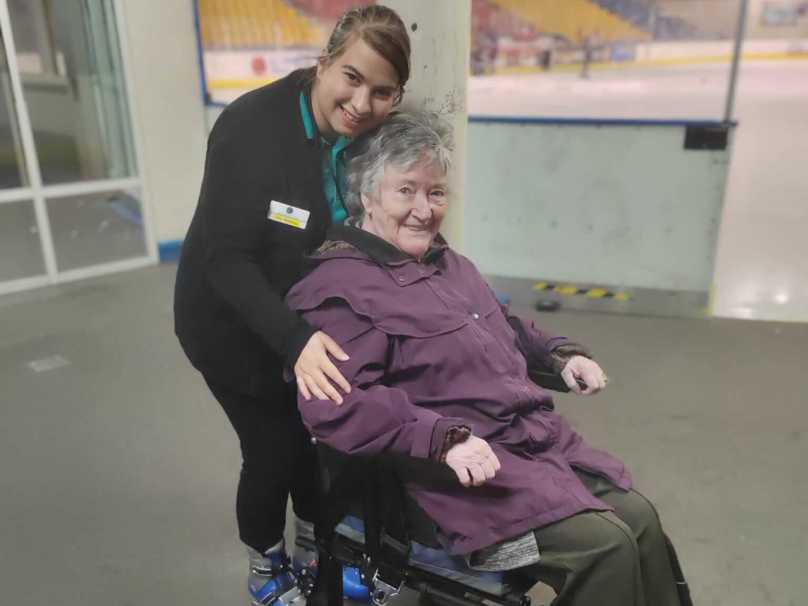 Staff member Ana stands behind resident Lynn, who is seated in a wheelchair eagerly awaiting her turn on the ice rink 