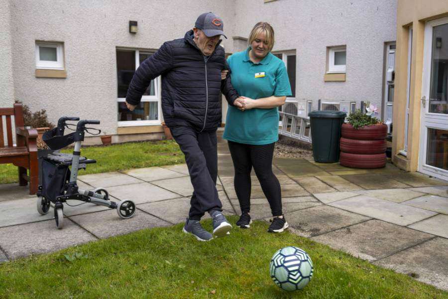 Care home resident stays active with a fun game of football assisted by a caring staff member outdoors