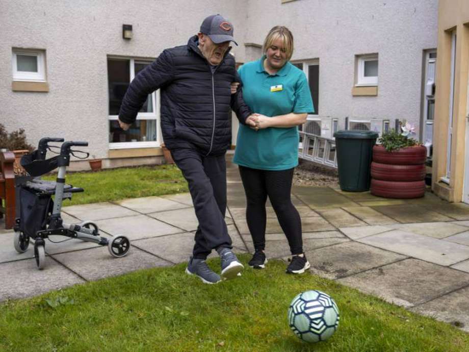 Care home resident stays active with a fun game of football assisted by a caring staff member outdoors