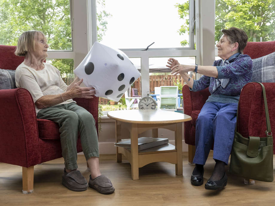 two woman are sitting in a communal lounge area on red armchairs, with a wooden table inbetween. The large windows at the back of them look out into the garden. They are throwing a large inflatable dice to eachother