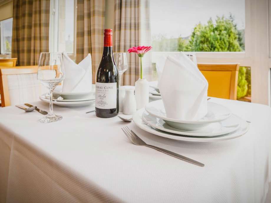 Decorative image of the dining table set up, focused on the tableware: napkins, plates, cutlery, a bottle of wine. Looking out the window to the garden space