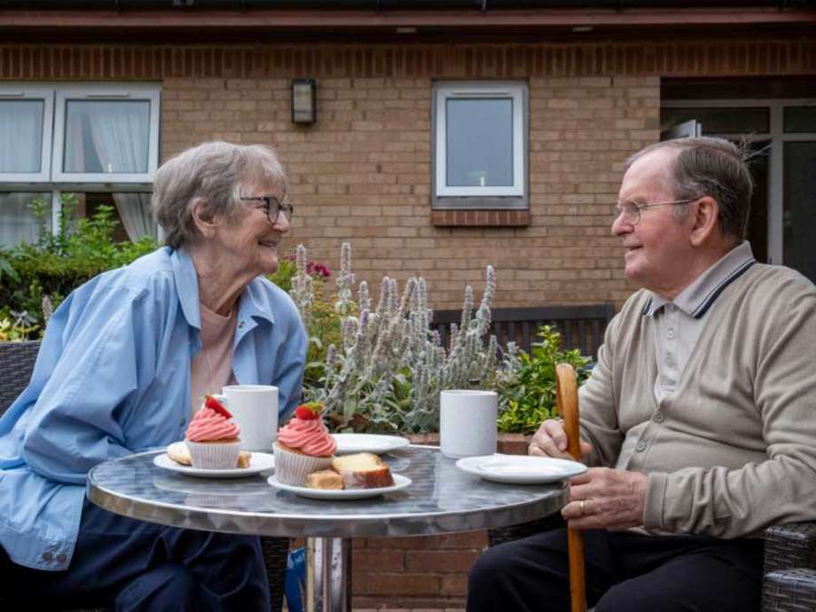 Two residents sit at an outdoor dining table set up for afternoon tea with cakes and teacups. They are smiling at one another and surrounded by plants with the care homes brick exterior in the background