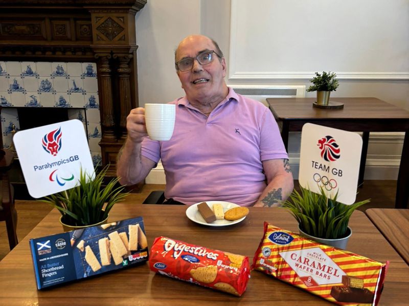 Care Home resident Ricky Tawse sits behind a table of biscuits holding up a cup of tea in search of britains most dunkable biscuit